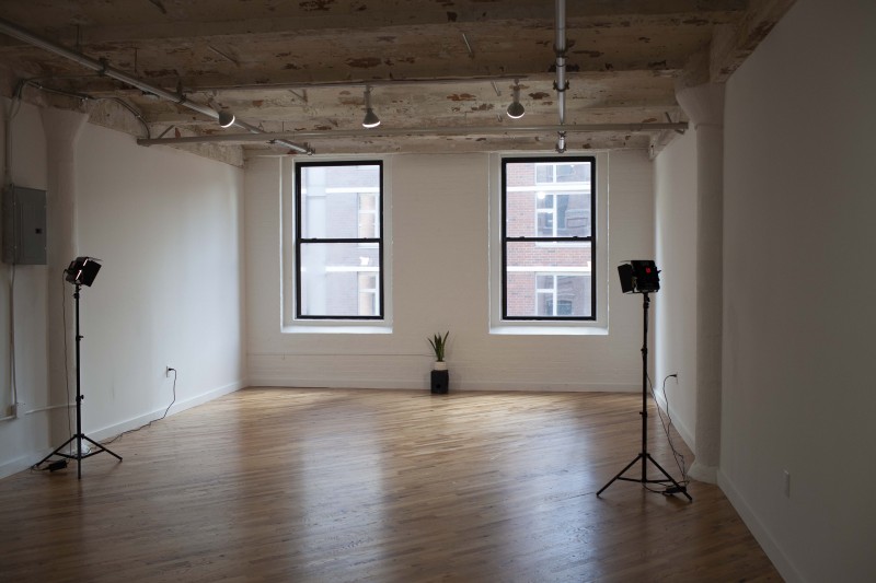 A sunlit studio with wooden floors and 2 windows. A small plant is on a speaker and 2 lights frame the space.