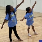 Two young dancers smile and pose with their arms extended in a bright dance studio 