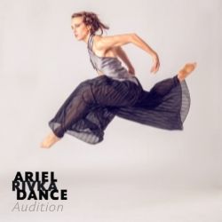 Woman jumping in air with legs split and arms bent back wearing purple pants and grey shirt