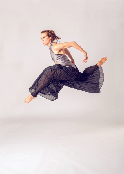 Woman in purple pants and grey top jumping in air with legs split and arms bent back