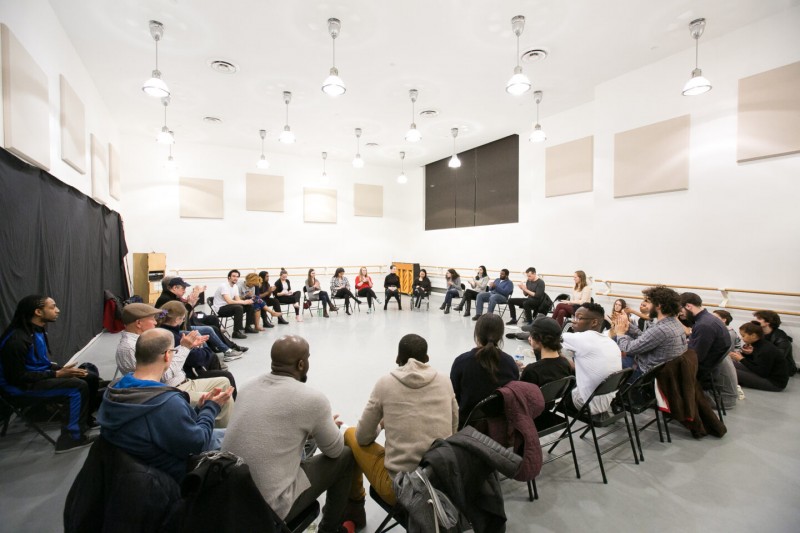 A group of people seated in chairs in a large circle that fills up the entire dance studio