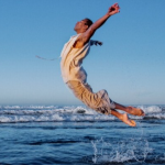 A dancer dressed in beige, jumps out of the ocean with his arms and legs slightly behind him creating a "C" shape in the air
