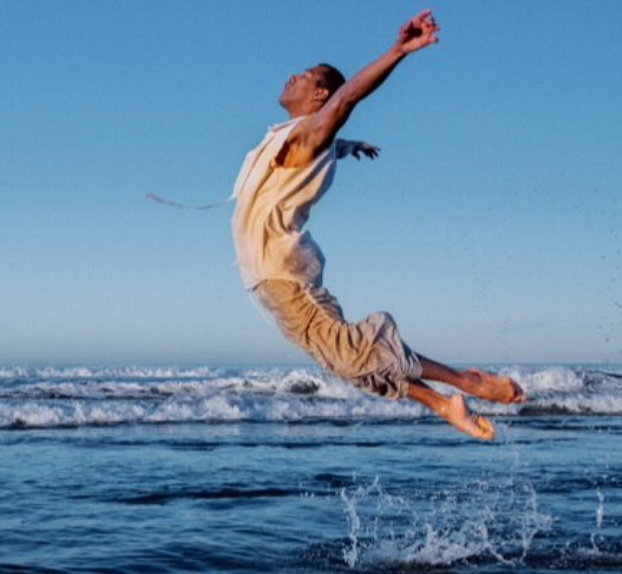 A dancer dressed in beige, jumps out of the ocean with his arms and legs slightly behind him creating a "C" shape in the air