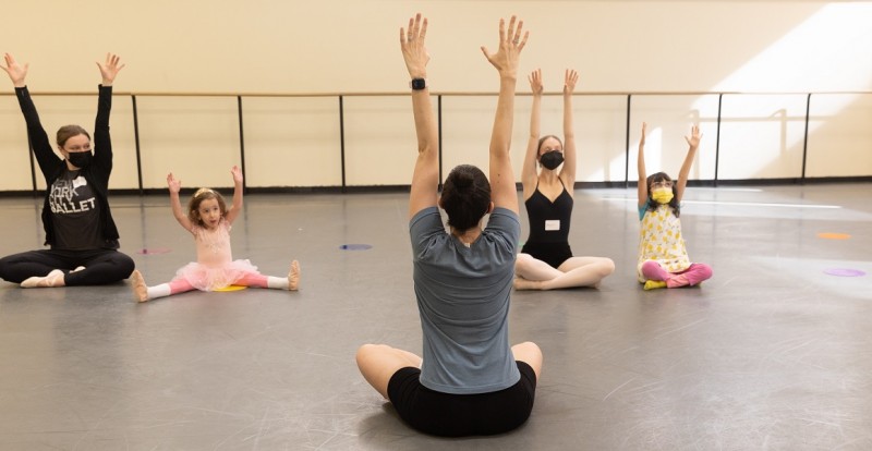 Children participating in NYCB Workshops sit on the ground with their arms stretched up into the air.