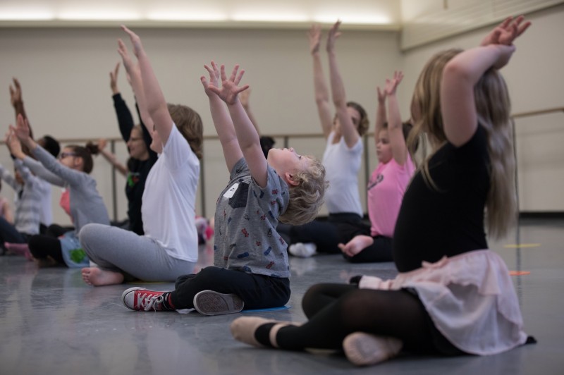 Children participating in NYCB Workshops sit on the ground with their arms stretched up into the air.