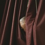 between the folds of a curtain a face in profile screams into the dark red fabric. The person is bald and asian.