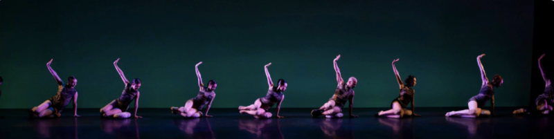 Company Dancers pictured in purple costumes on the floor of a stage with green and purple lighting
