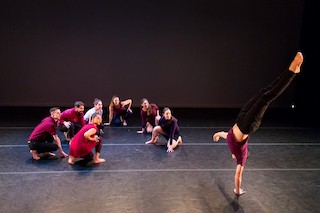 One dancer flips upside down as a cluster of dancers look on with excitement and joy. They are on stage with bright clothing.