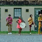 5 Music From The Sole dancers/ musicians jam in a line, in front of a farmhouse building.