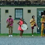 5 Music From The Sole dancers/ musicians jam in a line, in front of a farmhouse building.
