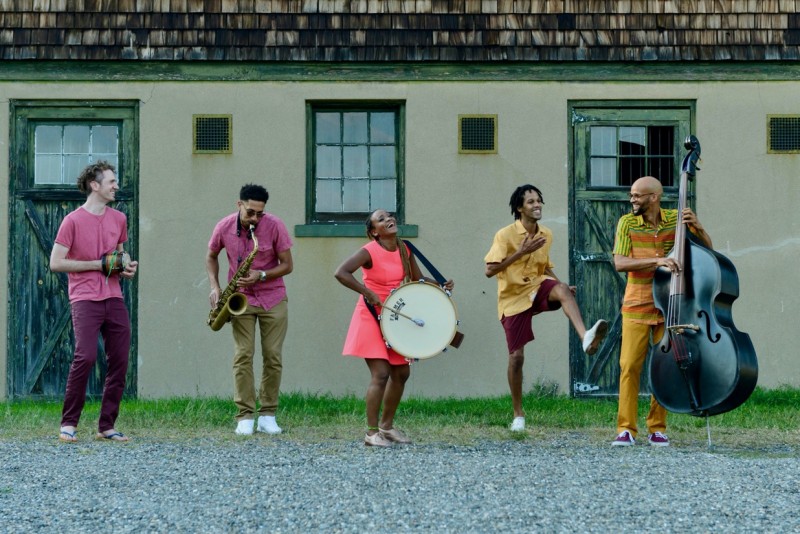 5 Music From The Sole dancers/ musicians jam in a line, in front of a farmhouse building.