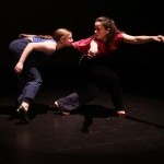 Two dancers on a dark stage - one biting the shirt of the other