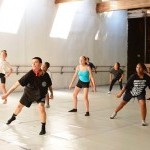 Sunlight falls through sky lights while a dance class takes place in a high-ceiling studio with a marley floor.