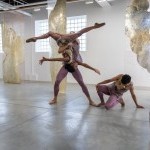 A female dancer is lifted in the air, upside down in the splits, as she reaching for a dancer kneeling on the floor.