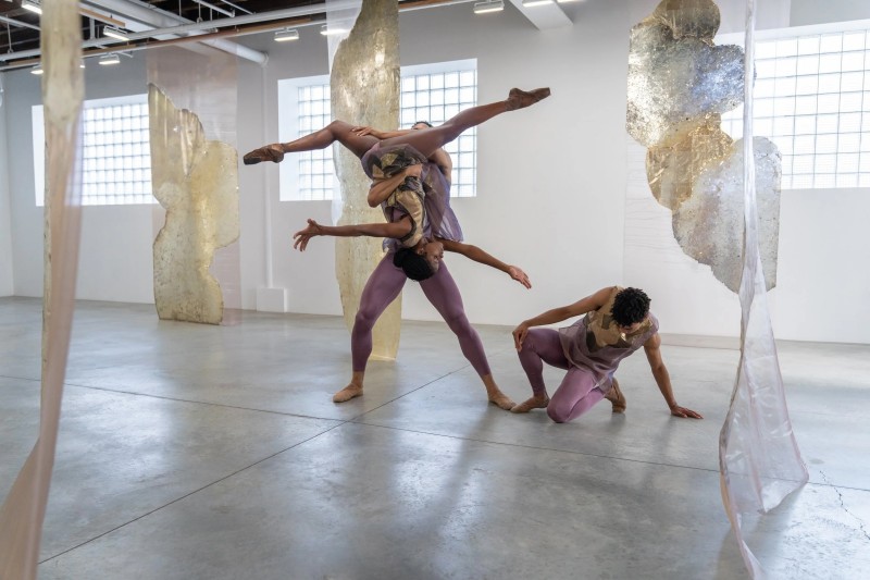 A female dancer is lifted in the air, upside down in the splits, as she reaching for a dancer kneeling on the floor.
