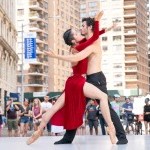 Two ballet dancers embrace on an outdoor city stage.