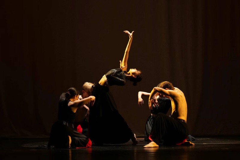 Dancers kneel in a circle around a soloist, who is standing in a backbend with her right arm gesturing to the sky.