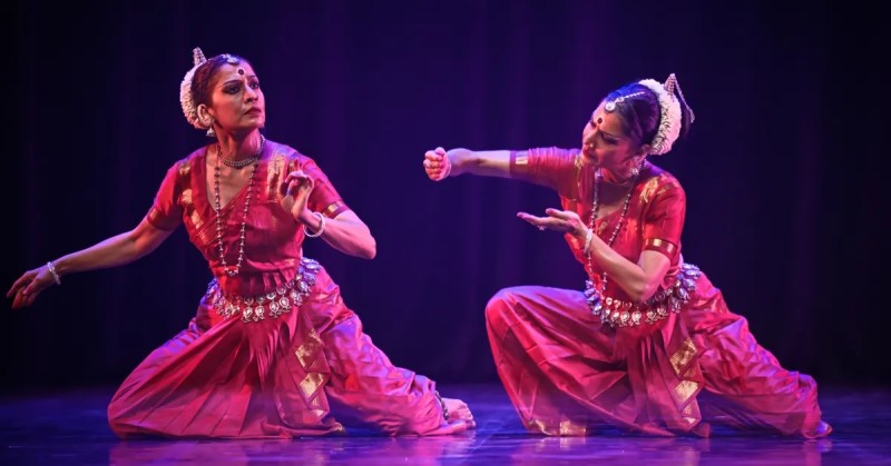 Two dancers perform a traditional Indian dance on stage, both dressed in vibrant red costumes adorned with silver jewelry.
