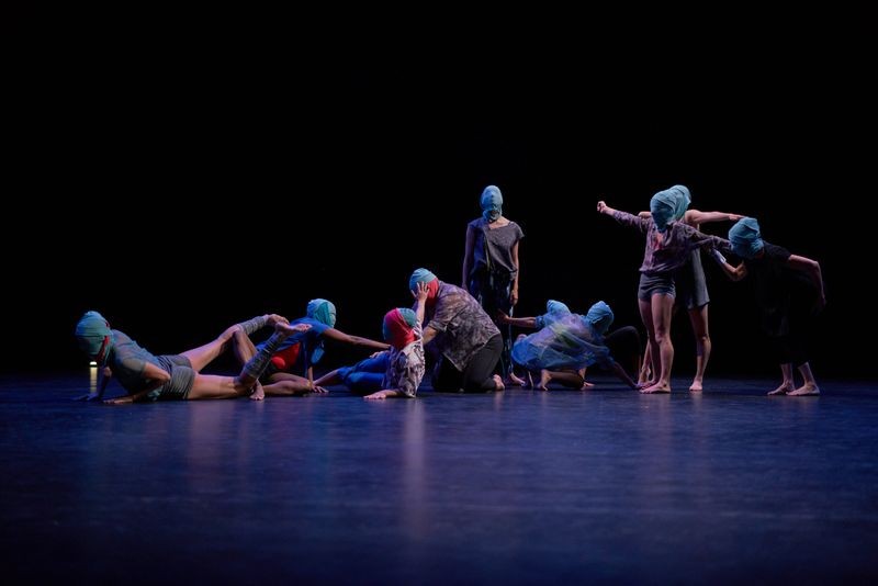 Ten dancers on stage, some on the ground and some standing, all with blue fabric wrapped around their faces and heads