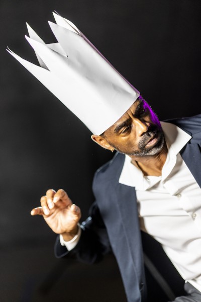 A Black man with facial hair is wearing a suit and a tall paper crown, he is looking off to the side