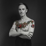 John Kelly as Samuel Steward, a shirtless white man with colorful tattoos wearing a hat and smoking a cigarette.