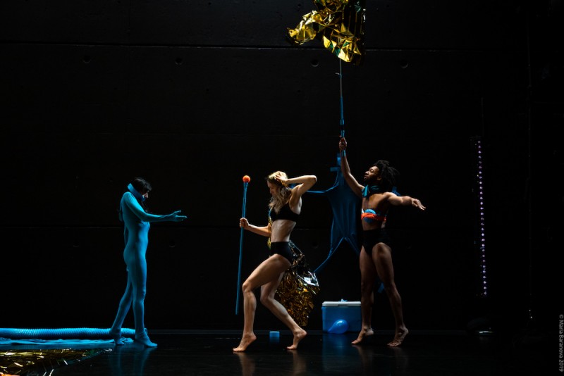 Three dancers on stage, one on the left is in a blue suit, two on the right are holding spears