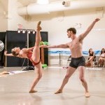 A woman in a reddish leotard leans backward lifting one leg skyward while being supported with one hand by a man in grey shorts