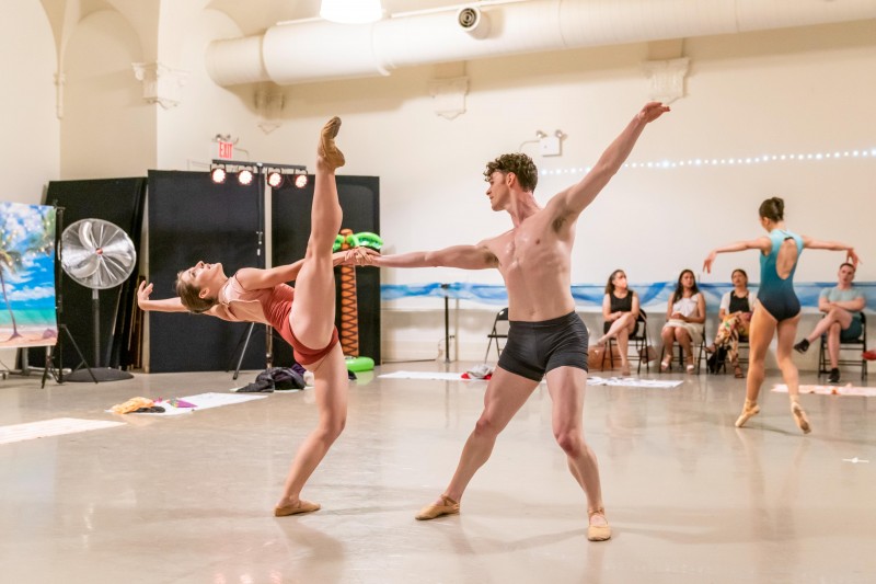 A woman in a reddish leotard leans backward lifting one leg skyward while being supported with one hand by a man in grey shorts
