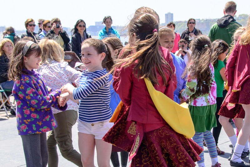 Image of children interlocking arms and dancing while an audience watches on in the background.