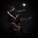 3 dancers in a row from top to bottom appear backlit from a single streetlamp