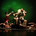 on stage a US Marine holds aloft his buddy surrounded by several dancers in black costumes.  A figure in red reaches towards him