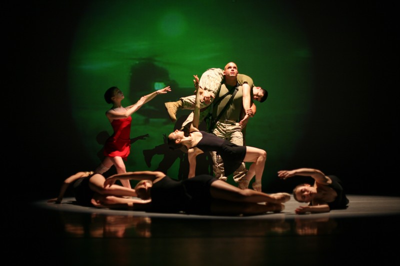 on stage a US Marine holds aloft his buddy surrounded by several dancers in black costumes.  A figure in red reaches towards him