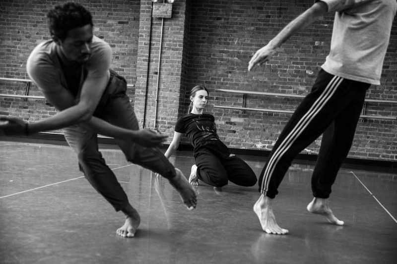 One dancer stooped over, falling to stage right, arms crossed, other dancer in the back spilling onto knees