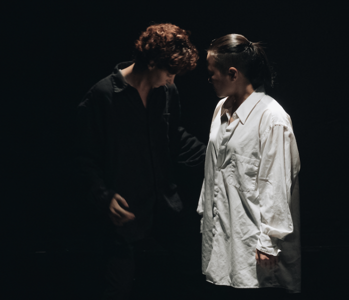 An intimate gesture featuring a dancer wearing black leaning into a dancer wearing white.