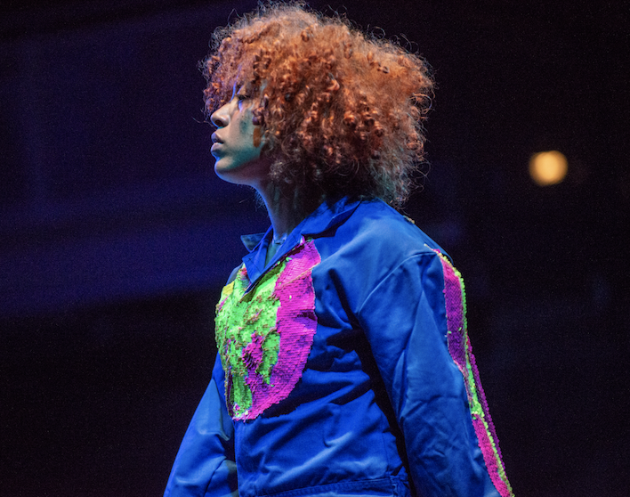 Dancer in profile, wearing a blue jumpsuit with appliqu&eacute;