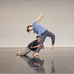 Two dancers against a gray background. One is low to the ground, leaning into the leg of the other.
