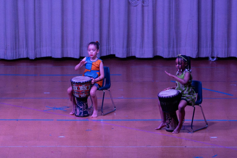 two young students wearing traditional african clothing sit in front of Djembe's on a stage