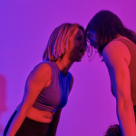 [image ID: In a pink and purple room, two women are screaming. Their arms are are rigid by their side as they tilt forward.}