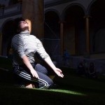 Dancer is kneeling in a spot of sunlight. Their focus is towards the sky.