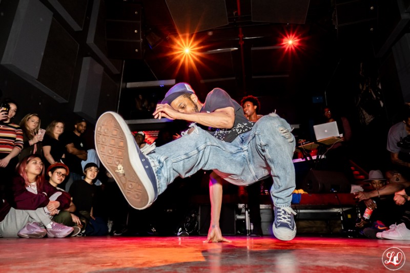 man dancing on the ground with a leg in the air in the foreground with audience watching in the background