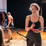 Brunette woman holding a notebook, smiling, talking to a musician with a dancer in the background.
