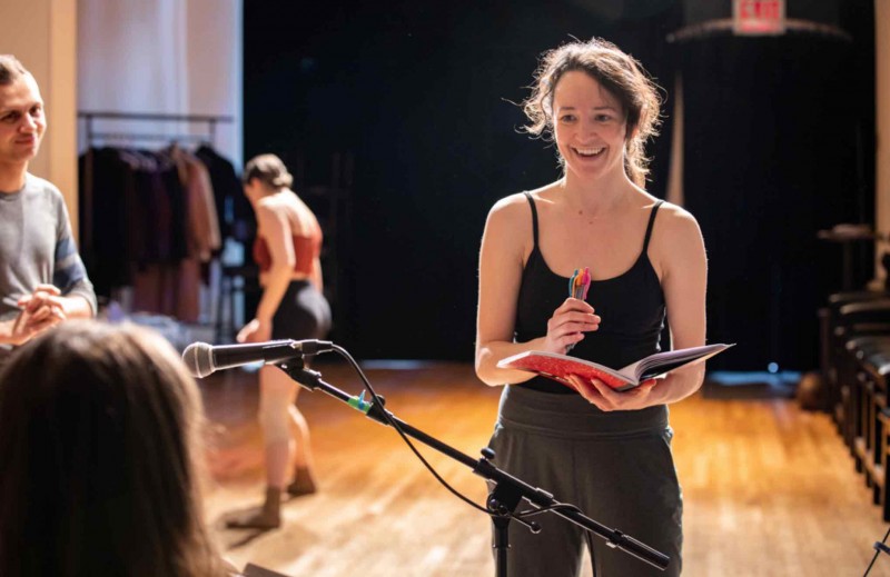 Brunette woman holding a notebook, smiling, talking to a musician with a dancer in the background. 