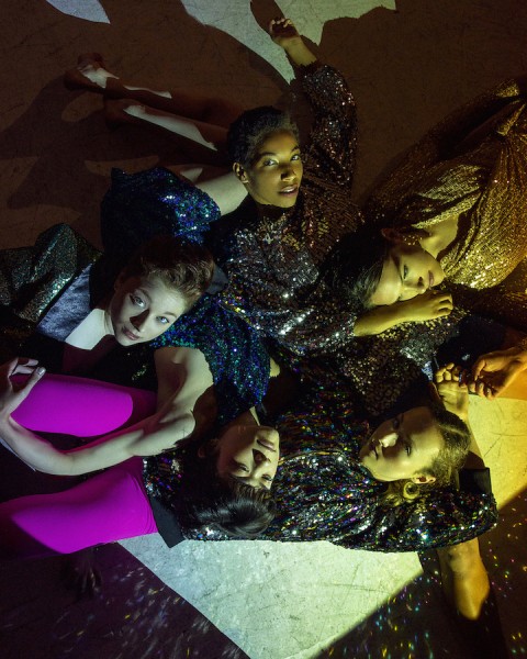 Five dance artists dressed in sequined outfits lay horizontally on the ground and their limbs pile atop one another