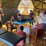 A spacious room with massage tables and hexagonally shaped windows. A group of people around massage tables watch a treatment.