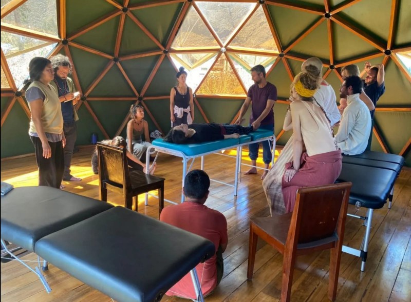 A spacious room with massage tables and hexagonally shaped windows. A group of people around massage tables watch a treatment.