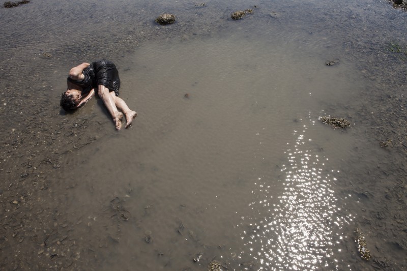 a woman lays, curled loosely around herself, in a thin layer of water on a grey mud flat. 