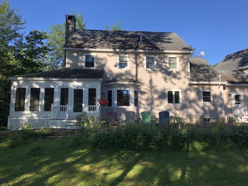 A pastel colored home sits tucked away behind a number of trees. Dappled sunlight hits the house and green grass of the backyard