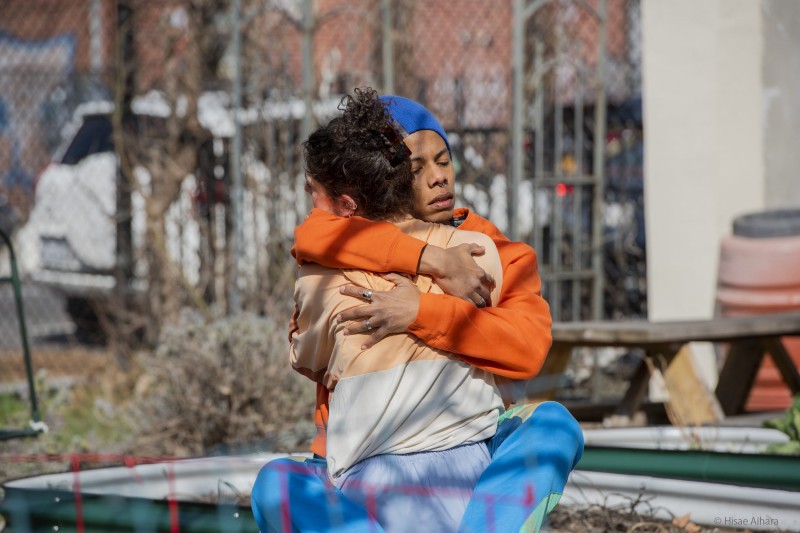 Photo represents two dancers in an embrace. Female identifying dancer is holding a male identifying dancer. They are outside.