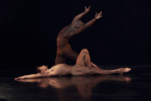 A dancer lays in his back; another dancer is bent over him, arms raised to the air in a dramatic flourish