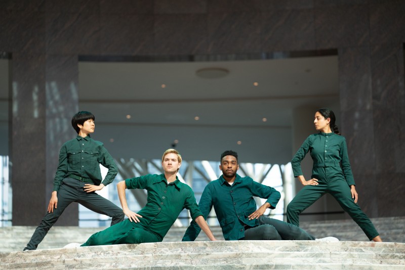 four dancers in green coveralls pose at the top of a white marble staircase, forming a statuesque, symmetrical shape